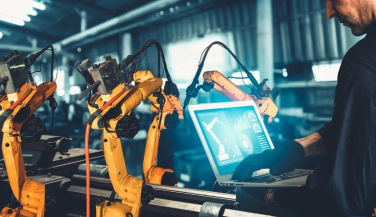 A worker uses a laptop to control a set of robotic arms to represent industrial automationl.